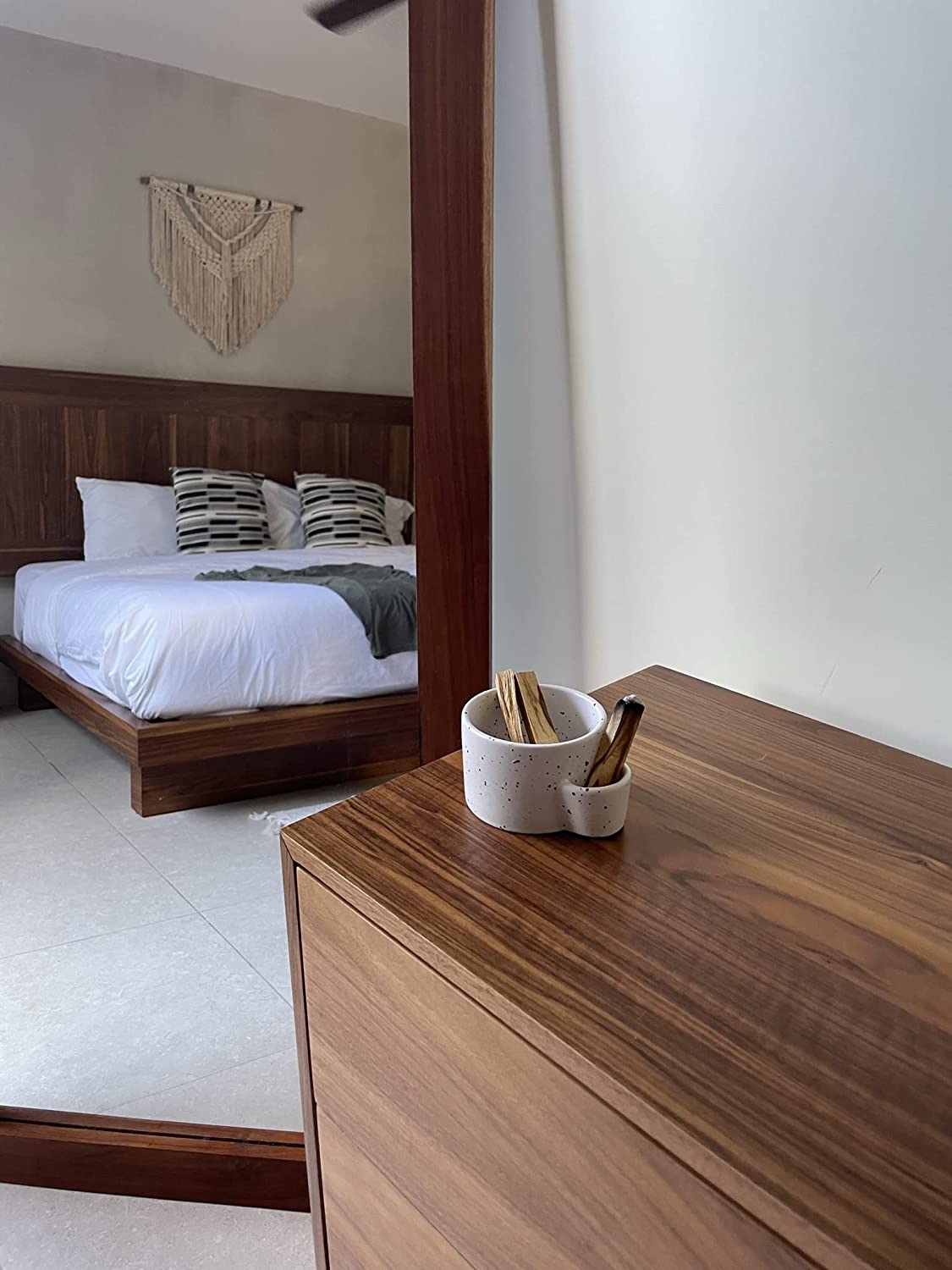 Wooden dresser with a mirror reflecting a bedroom with a bed and decorative wall hanging.