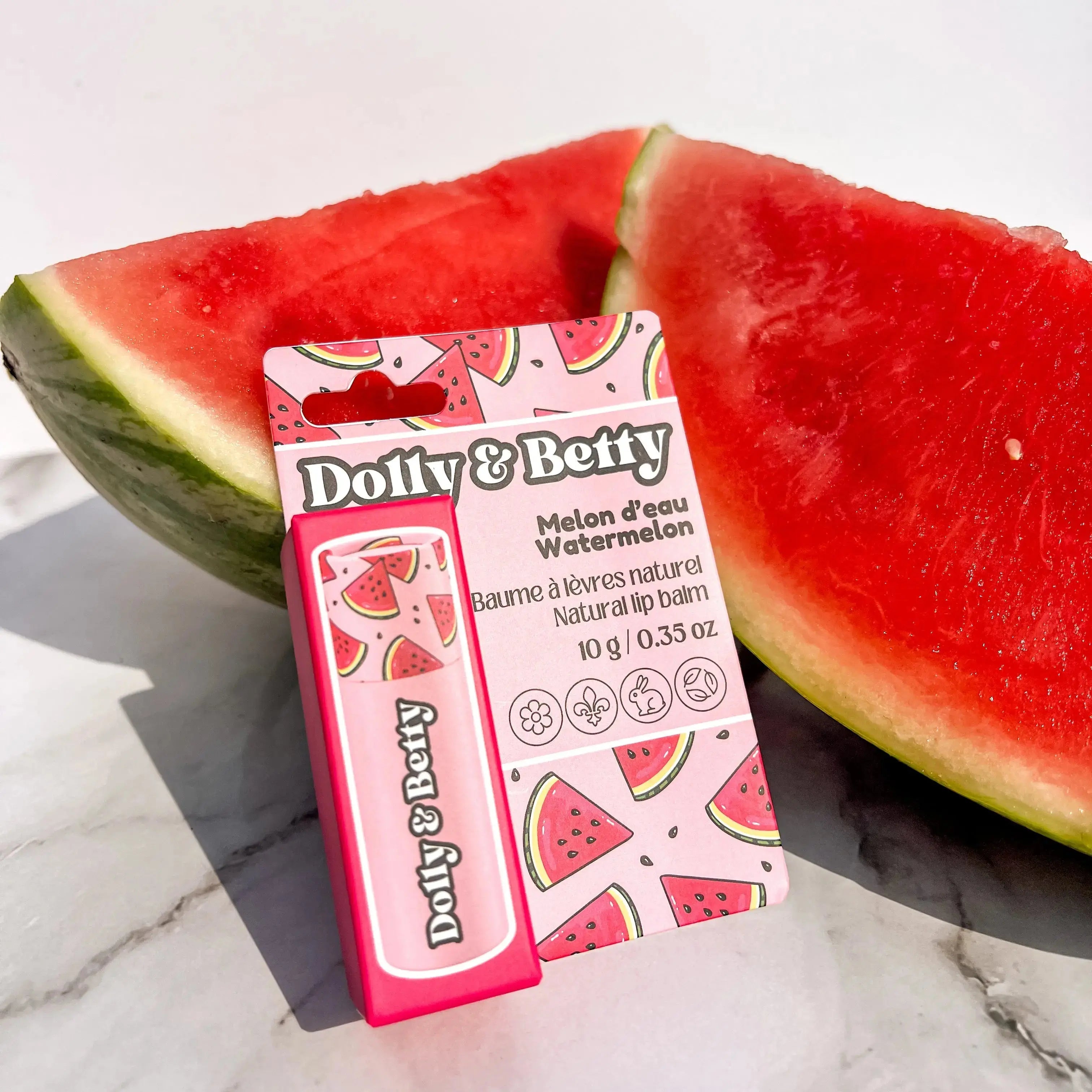 Dotty & Betty lip balm packaging with watermelon design next to a slice of watermelon on a marble surface