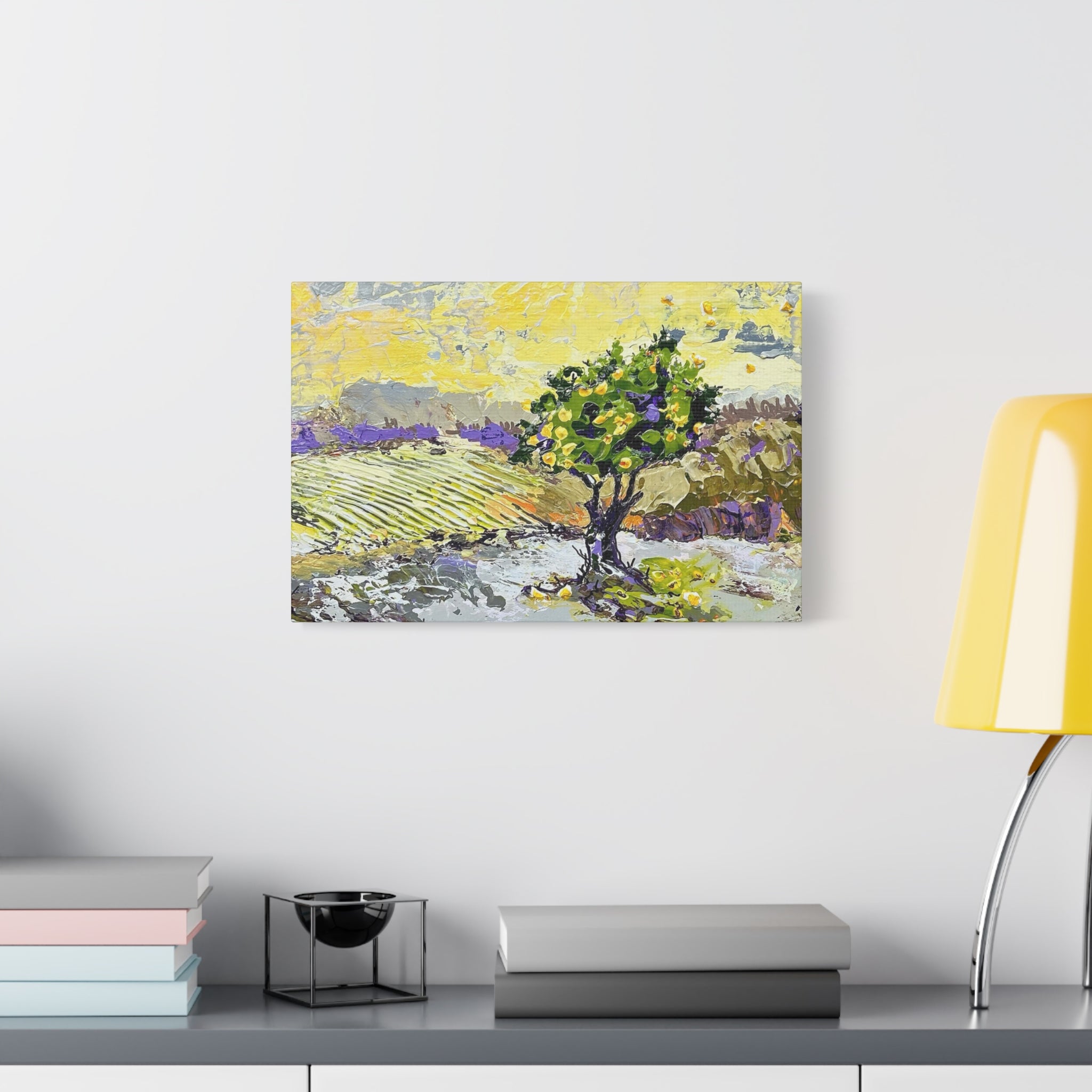 A square canvas art print depicting a landscape with a green tree and purple hills under a yellow sky, displayed on a white wall above a gray console table with books and a yellow lamp.