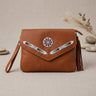 OldTribes A brown leather envelope-style handbag with a wrist strap, tassel detail, and colorful beaded geometric accents on the front flap, displayed on a neutral textured beige surface with decorative stones and dried grass in the background.