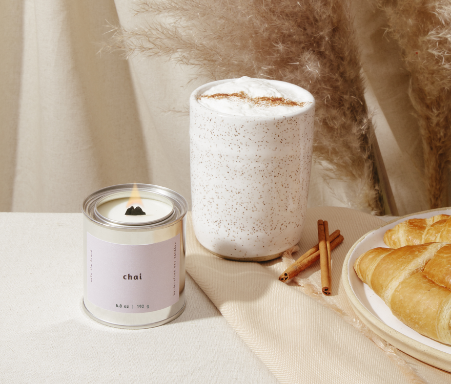 Candle labeled 'chai' next to a cup of coffee and pastries on a table.