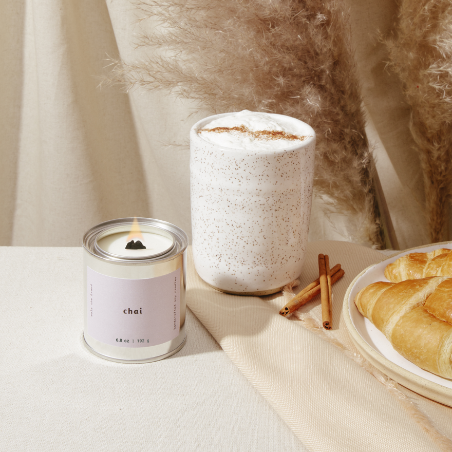 Candle labeled 'chai' next to a cup of coffee and pastries on a table.