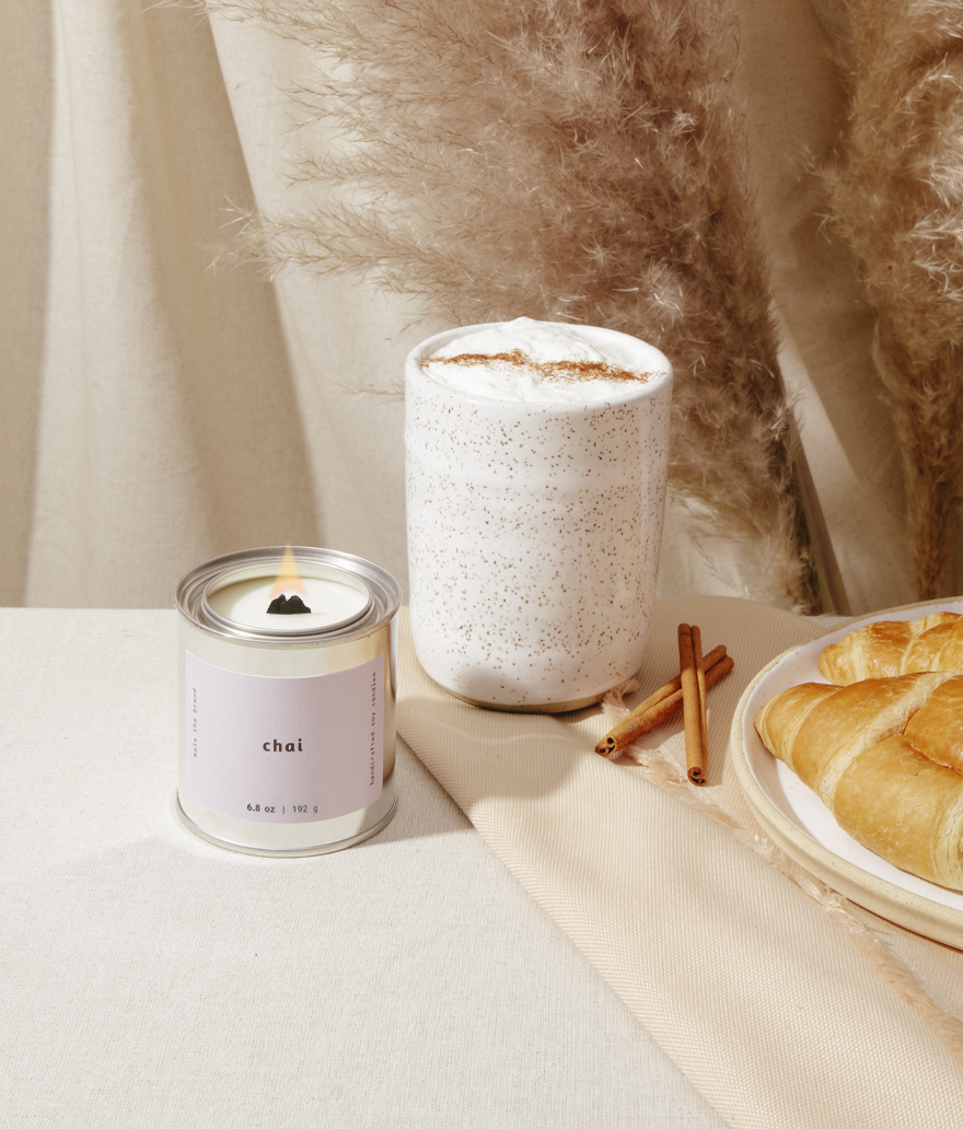 Candle labeled 'chai' next to a cup of coffee and pastries on a table.
