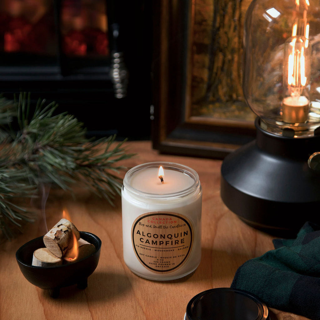 Candle labeled 'Alpena Quill Campfire' on a wooden surface with a fireplace and lantern in the background.