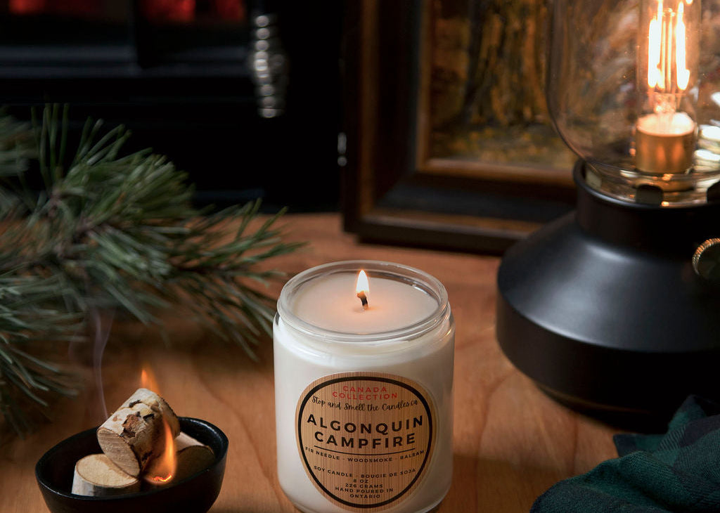 Candle labeled 'Alpena Quill Campfire' on a wooden surface with a fireplace and lantern in the background.
