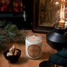 Candle labeled 'Alpena Quill Campfire' on a wooden surface with a fireplace and lantern in the background.