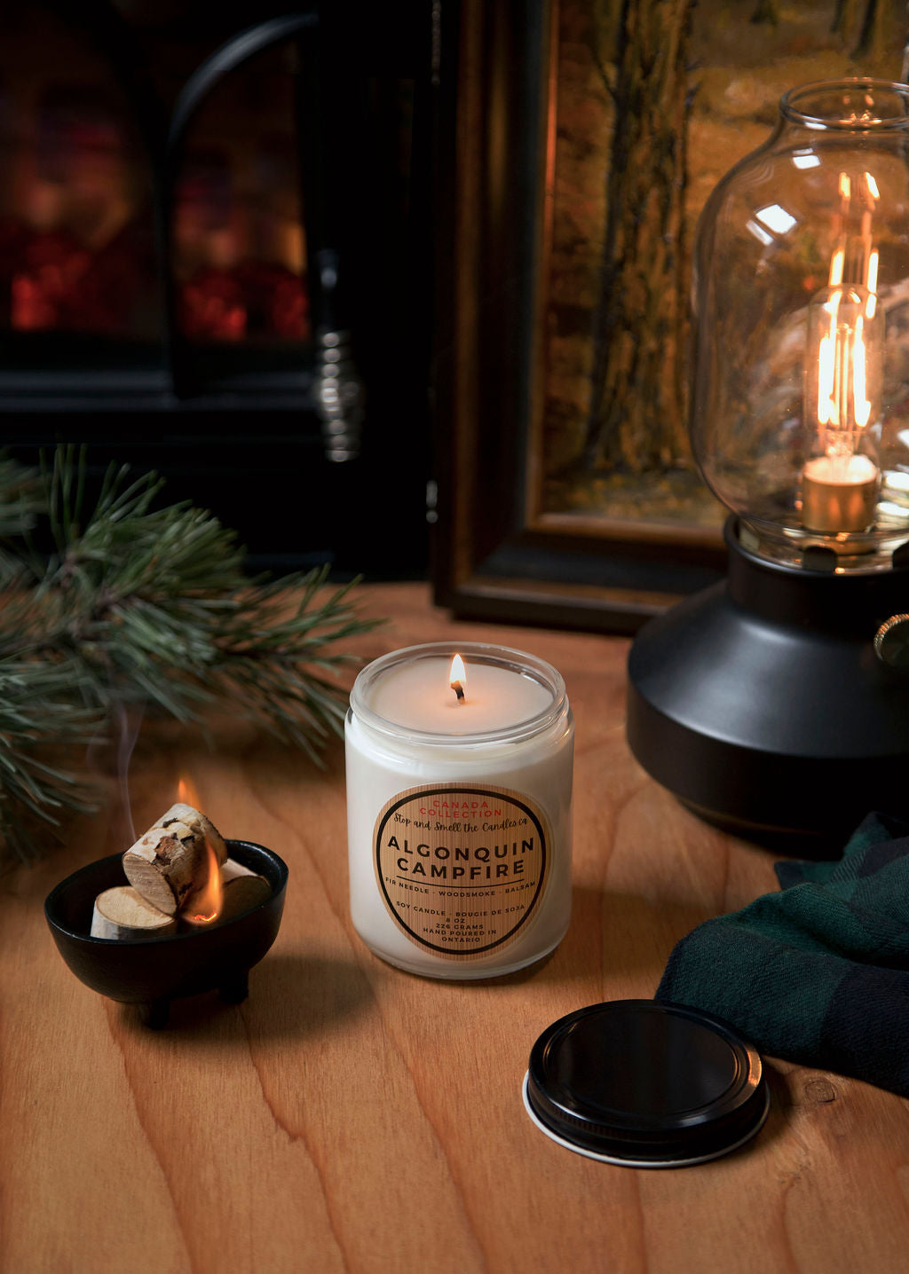Candle labeled 'Alpena Quill Campfire' on a wooden surface with a fireplace and lantern in the background.