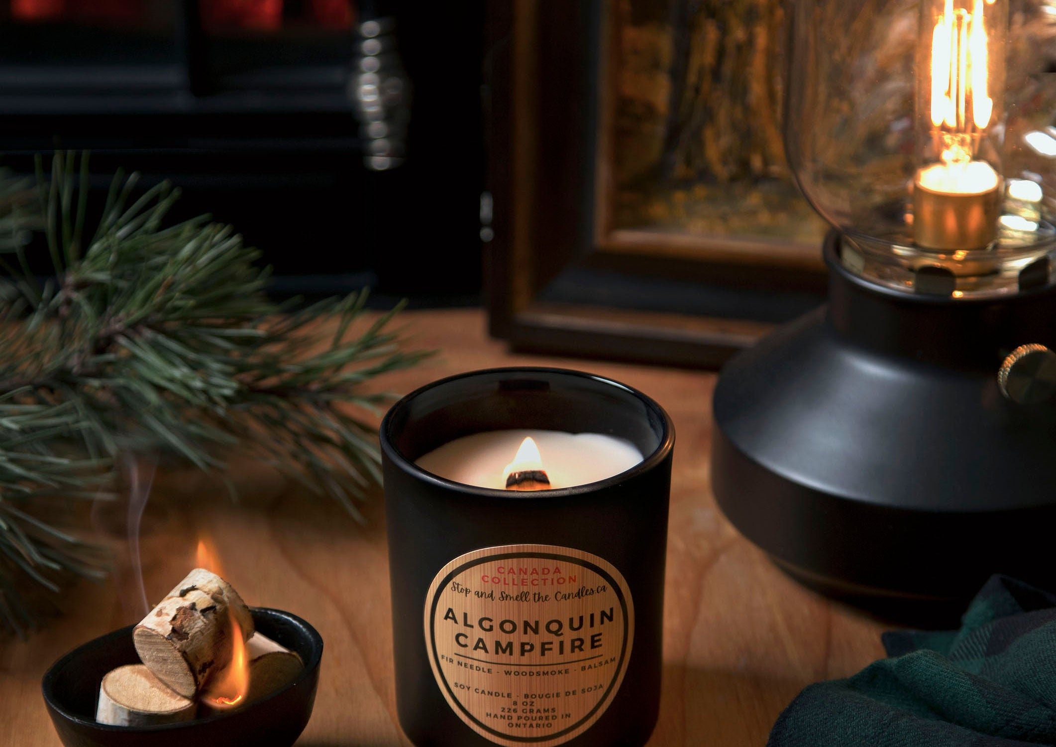Stopandsmellthecandles Algonquin Campfire soy candle – 8 oz black jar with lit wooden wick and wooden lid beside pine branches