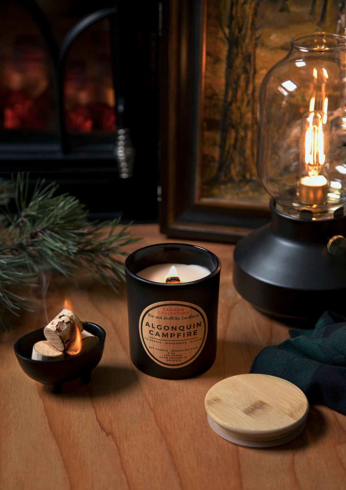 Stopandsmellthecandles Algonquin Campfire soy candle – 8 oz black jar with lit wooden wick and wooden lid beside pine branches