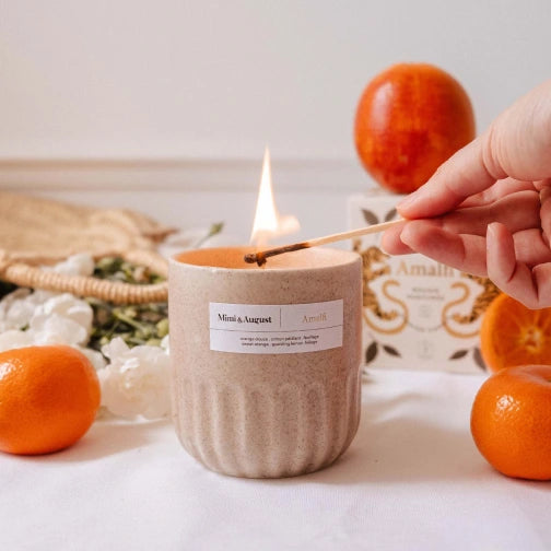 Amalfi candle with citrus and tropical scent, Candle being lit with a match, surrounded by oranges and a woven basket on a light surface.