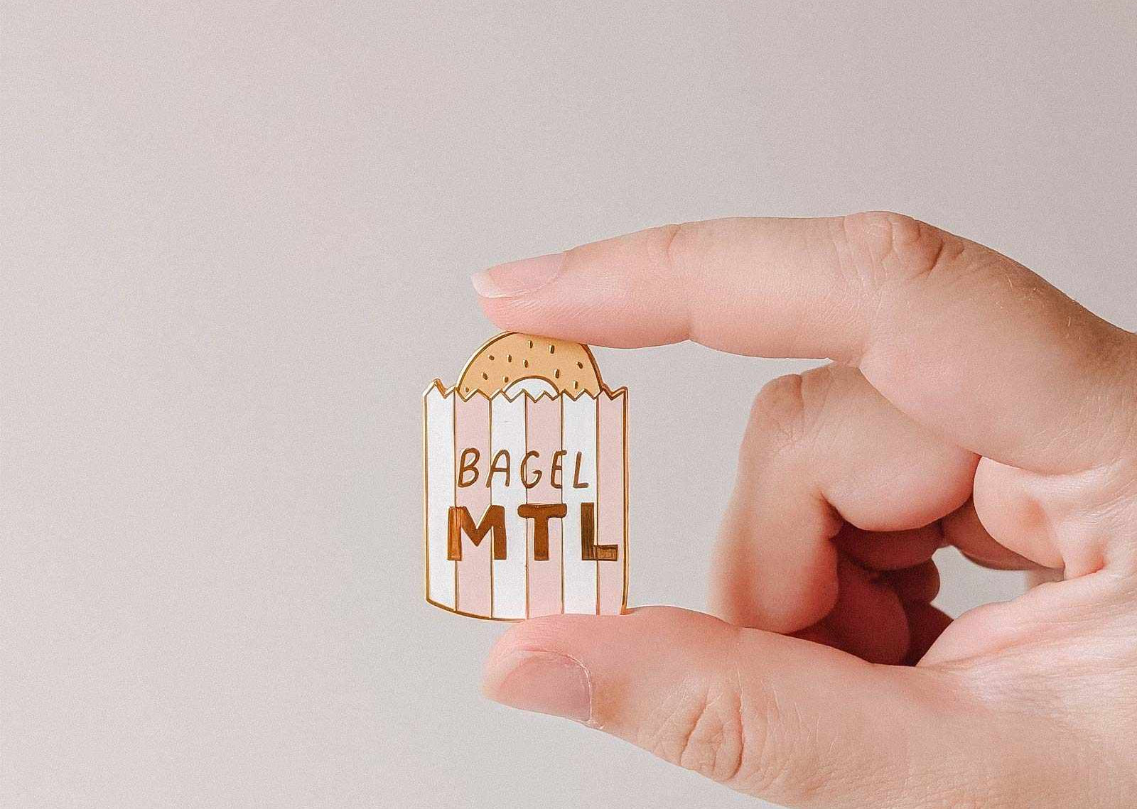 mimi-and-august Hand holding a bagel-shaped enamel pin with 'Bagel MTL' text on a plain background