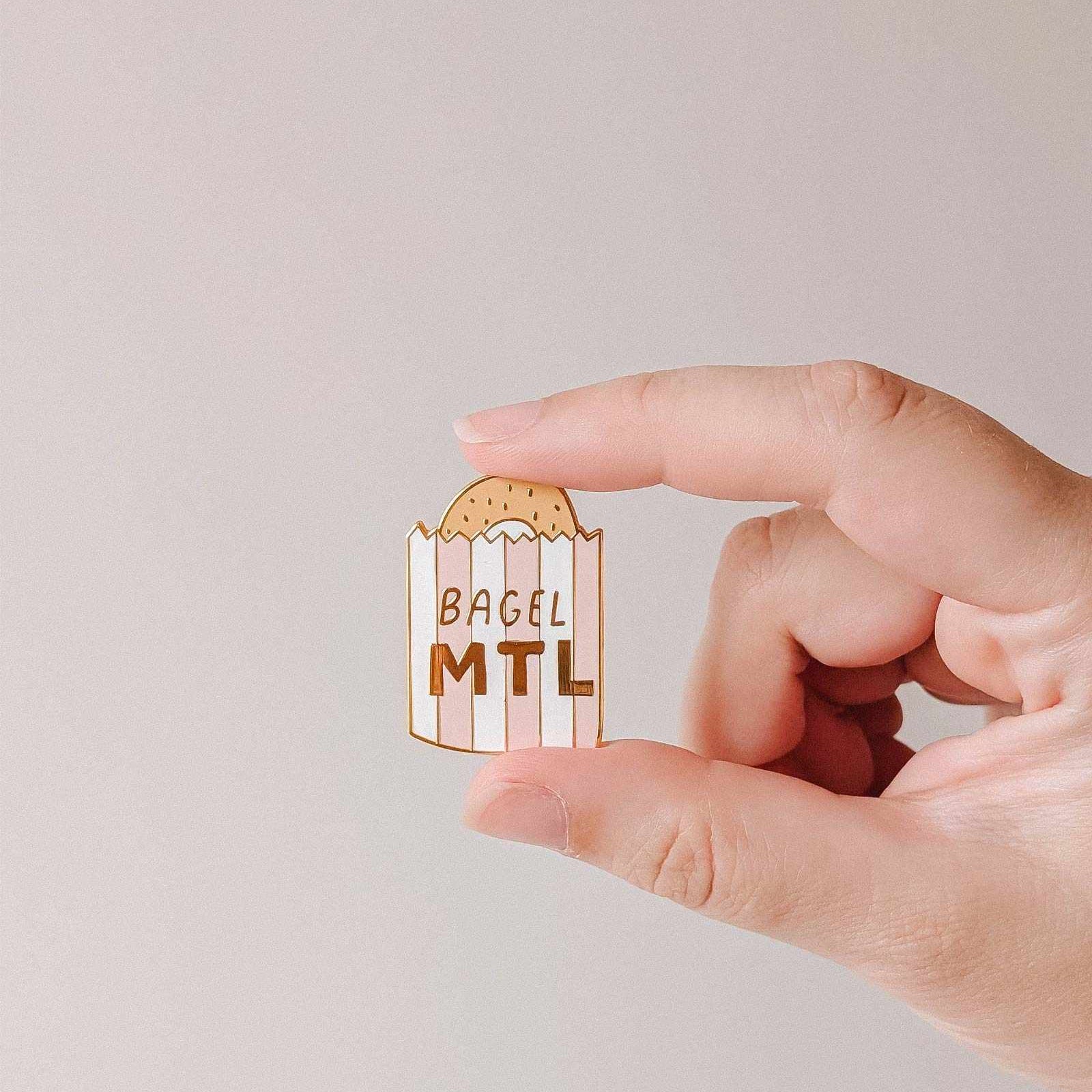 mimi-and-august Hand holding a bagel-shaped enamel pin with 'Bagel MTL' text on a plain background