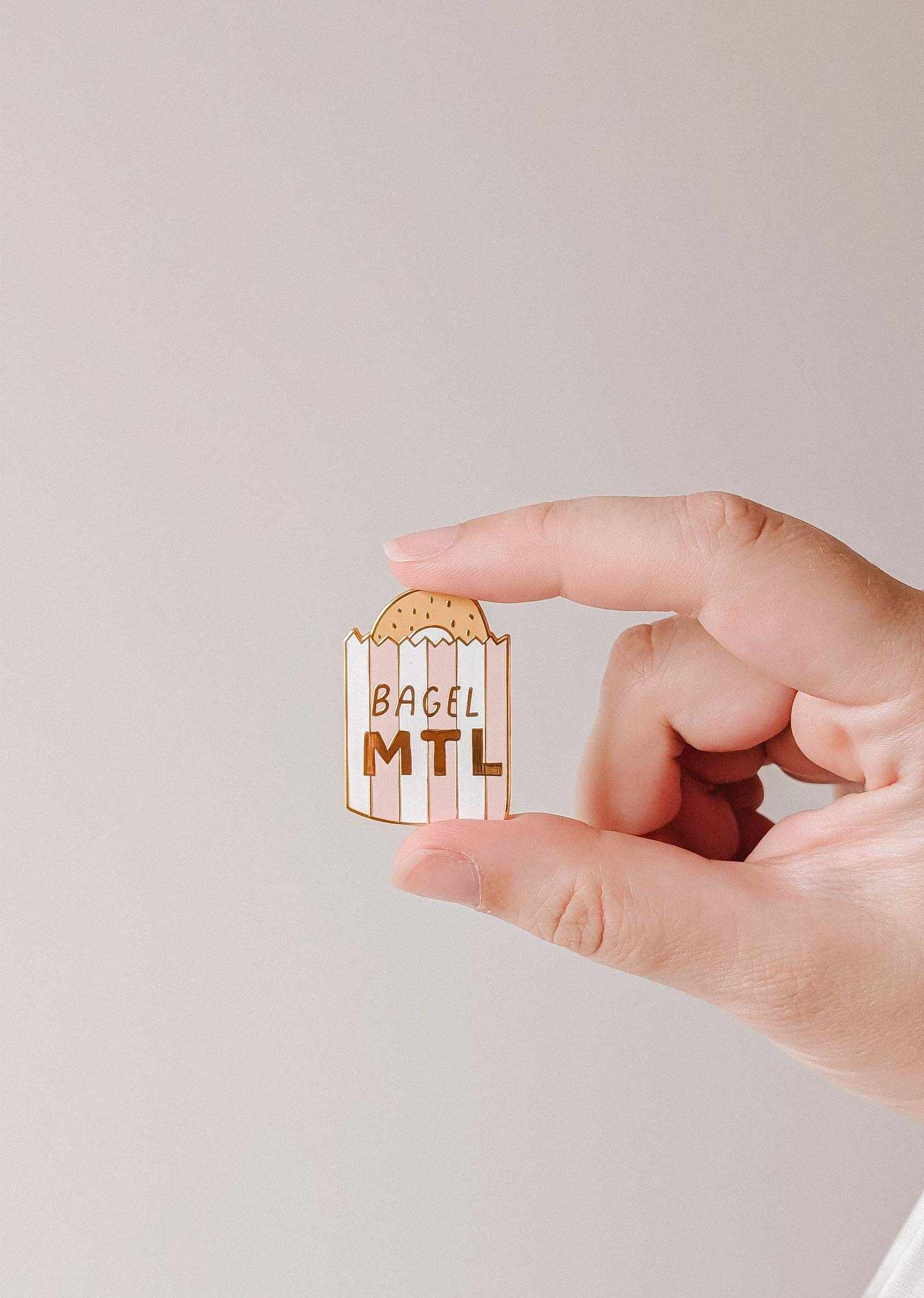 mimi-and-august Hand holding a bagel-shaped enamel pin with 'Bagel MTL' text on a plain background