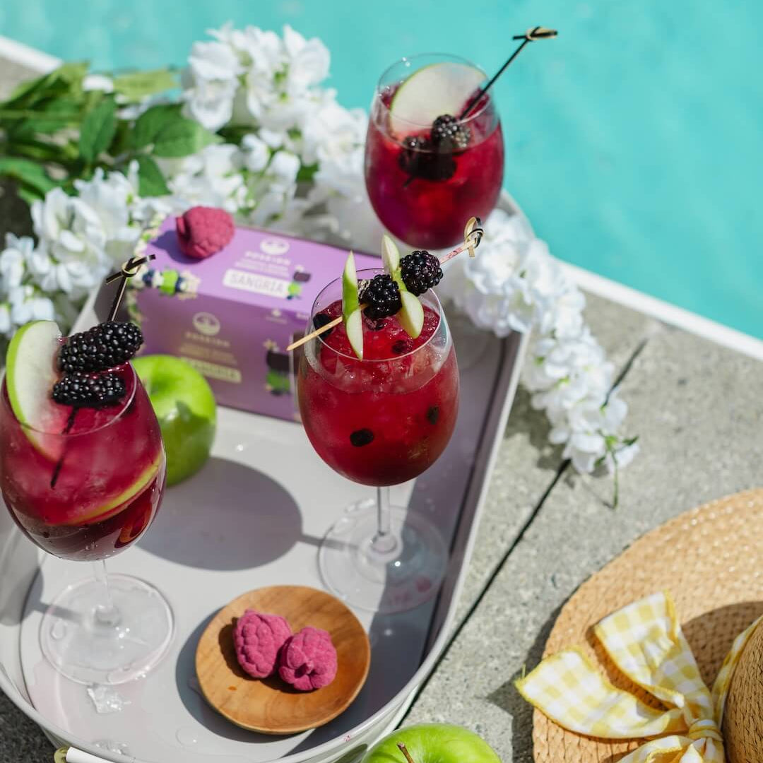 Three red mocktails with fruit flavor bombs on a tray by a poolside, surrounded by apples and flowers.