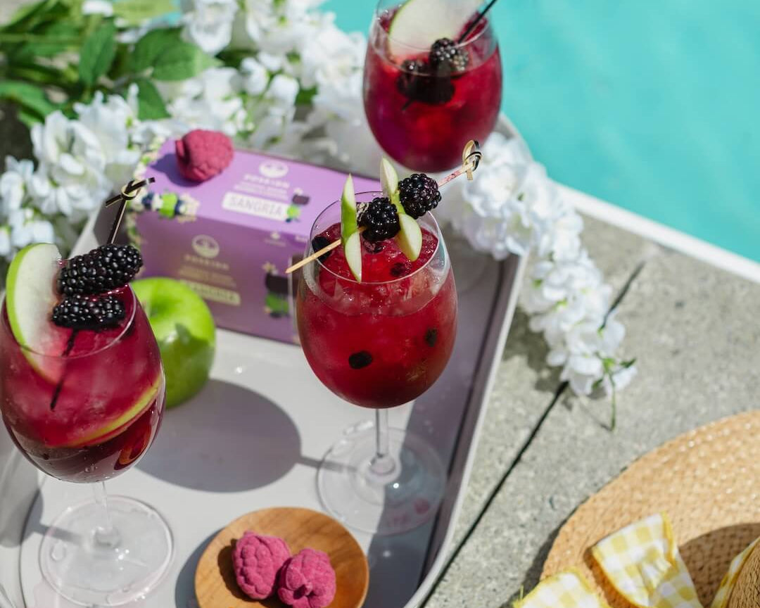 Three red mocktails with fruit flavor bombs on a tray by a poolside, surrounded by apples and flowers.