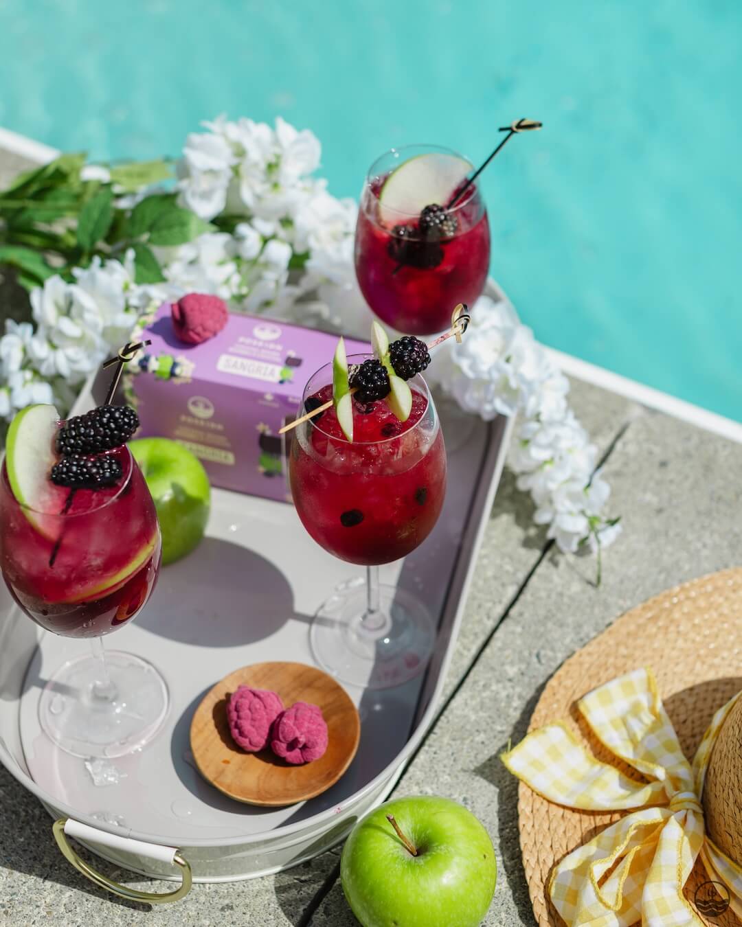 Three red mocktails with fruit flavor bombs on a tray by a poolside, surrounded by apples and flowers.