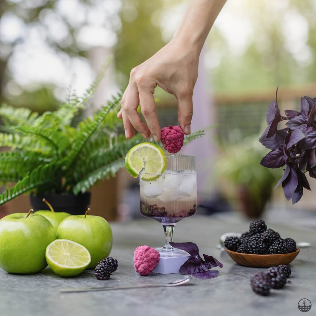 Hand placing a pink fruit flavor bomb on a mocktail garnished with lime and ice, surrounded by apples, limes, and blackberries on a table.