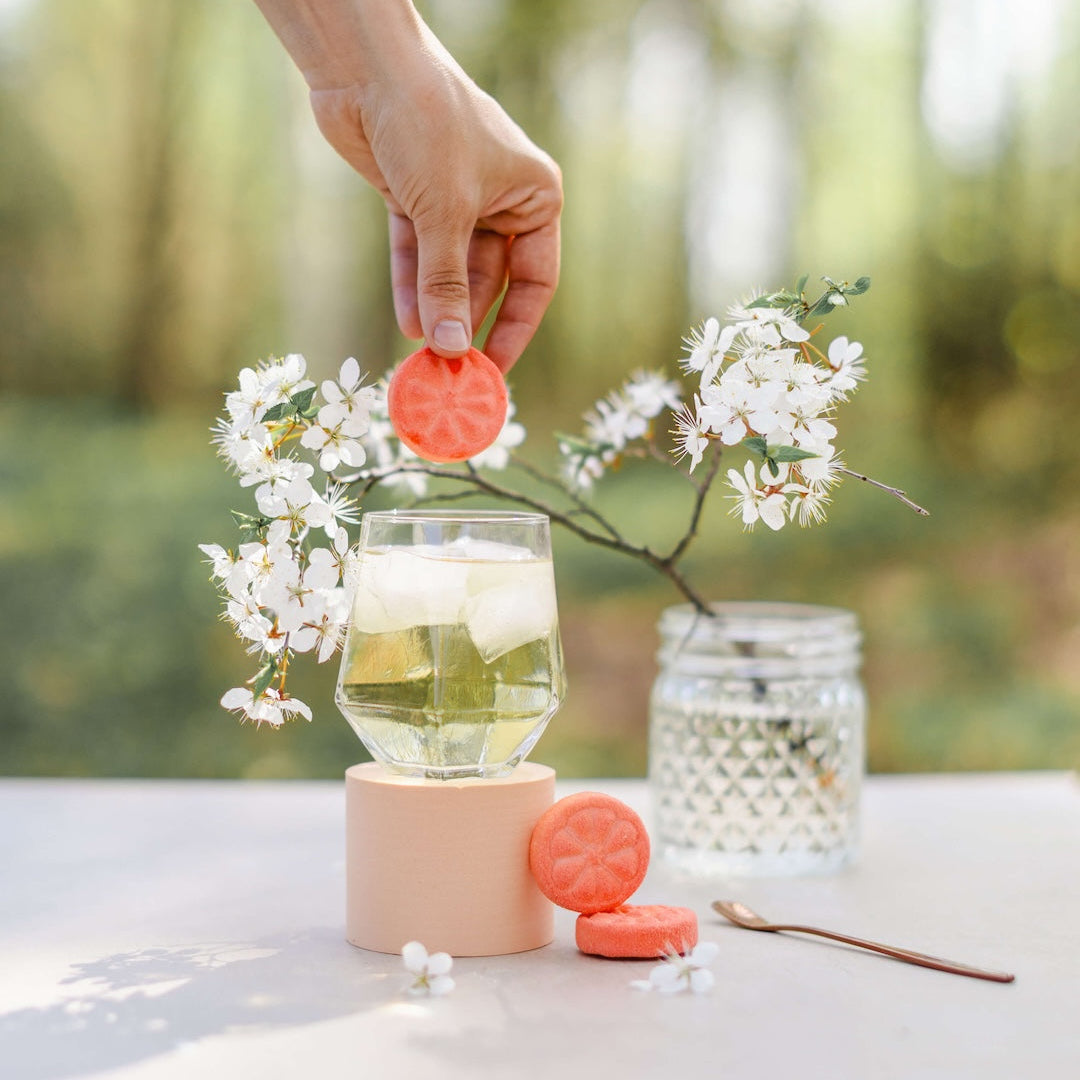 Person holding a pink lid over a glass of lemonade with flowers and a candle on a table outdoors. Poseidn