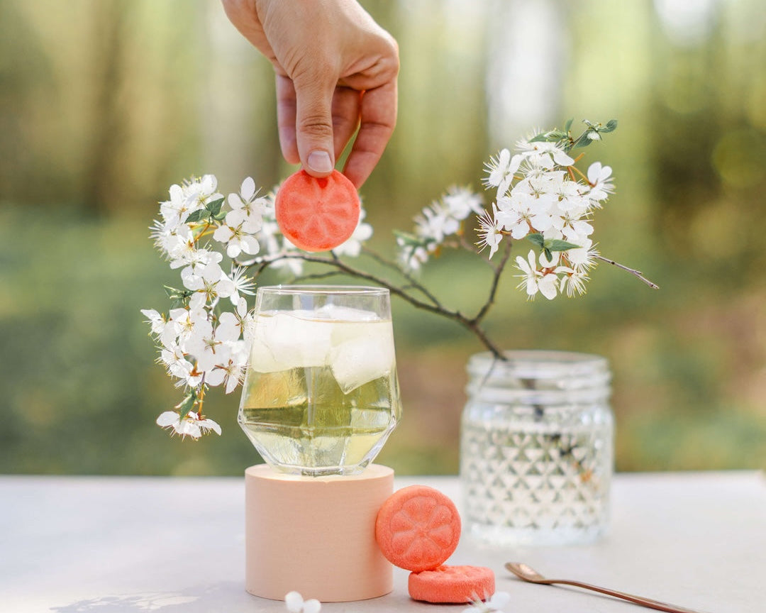 Person holding a pink lid over a glass of lemonade with flowers and a candle on a table outdoors. Poseidn