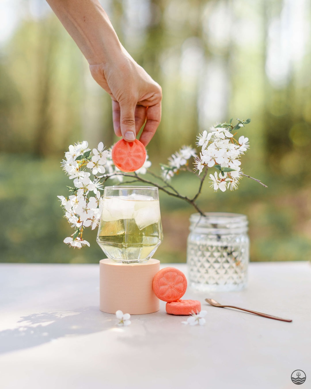 Person holding a pink lid over a glass of lemonade with flowers and a candle on a table outdoors. Poseidn