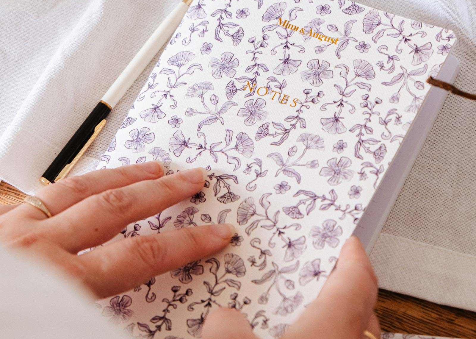 mimi-and-august Person holding a floral notebook with a pen on a light surface