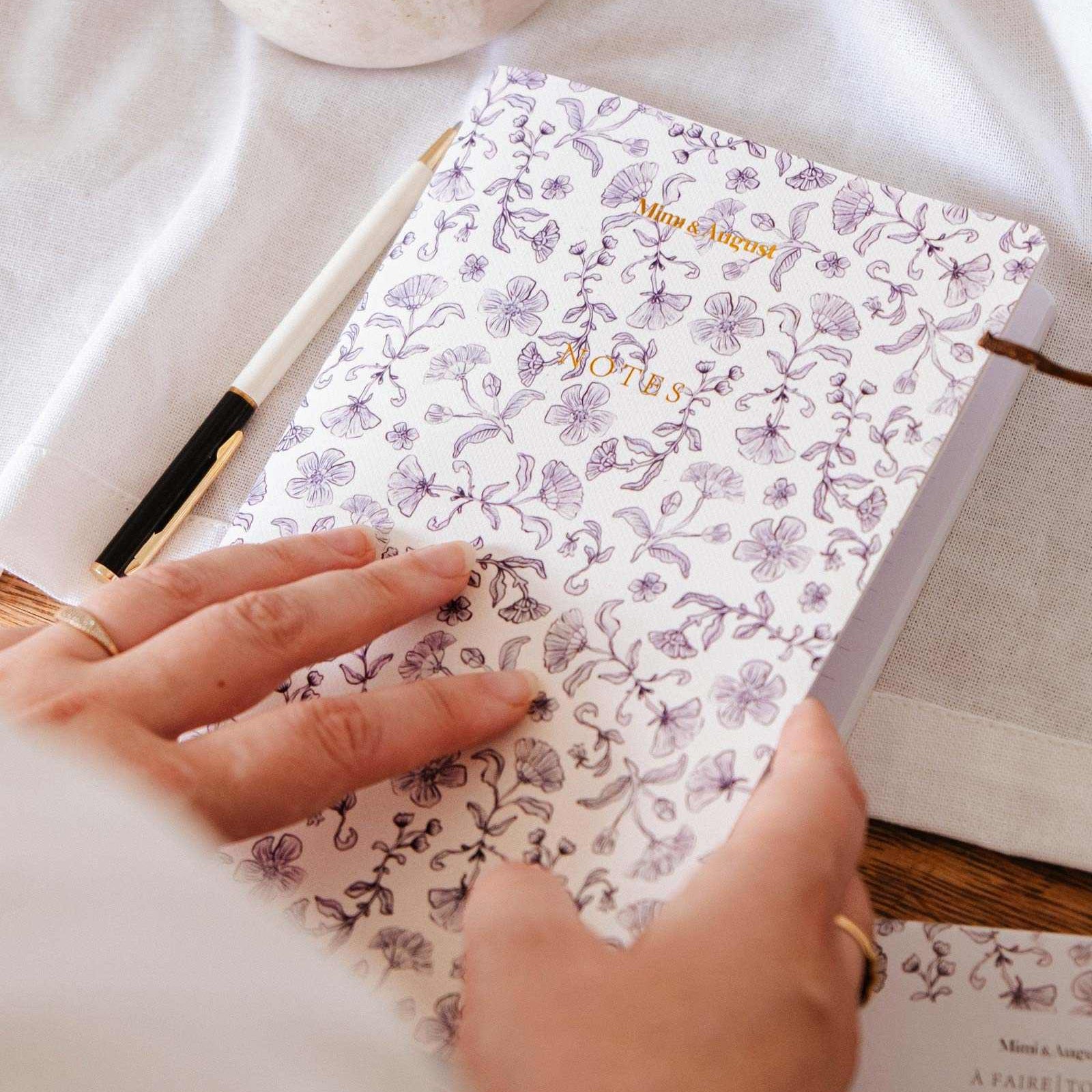 mimi-and-august Person holding a floral notebook with a pen on a light surface