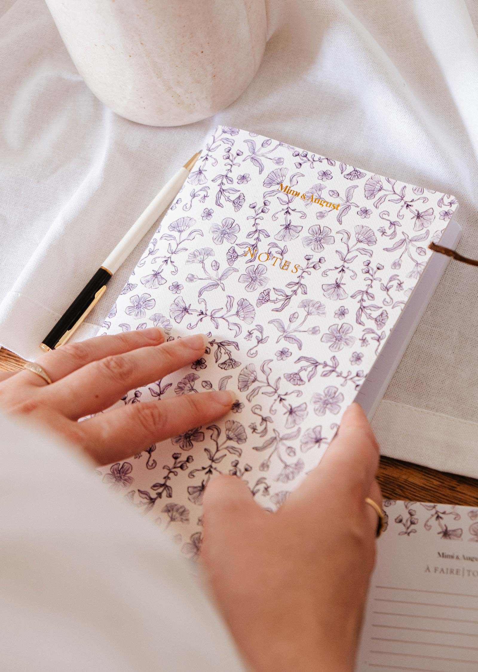 mimi-and-august Person holding a floral notebook with a pen on a light surface
