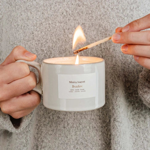 Person lighting a candle in a white mug labeled Bonfire Reusable Candle by mimi and august