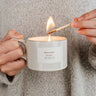 Person lighting a candle in a white mug labeled Bonfire Reusable Candle by mimi and august