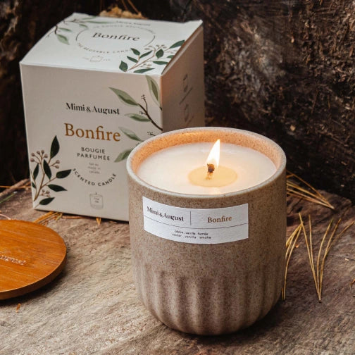 Mimi & August Bonfire candle with packaging on a wooden surface Person lighting a candle in a white mug labeled Bonfire Reusable Candle by mimi and august
