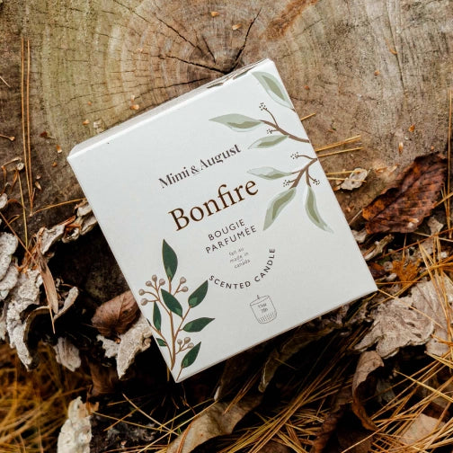 Mimi & August Bonfire scented candle packaging on a wooden surface with leaves and twigs.