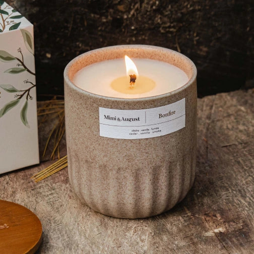 Ceramic candle with a label on a wooden surface Person lighting a candle in a white mug labeled Bonfire Reusable Candle by mimi and august