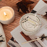 Candle with a label on top of a magazine on a wooden surface a candle in a ceramic mug labeled Bonfire Reusable Candle by mimi and august