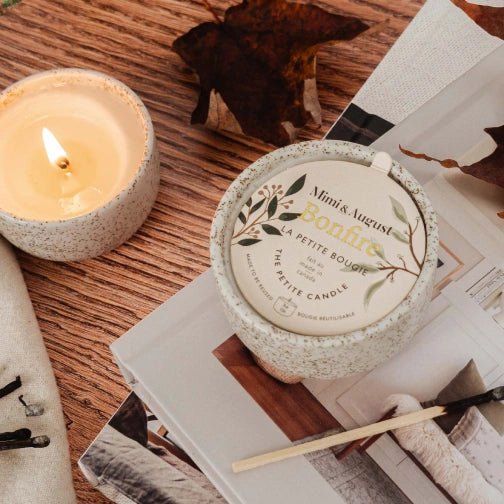 Candle with a label on top of a magazine on a wooden surface a candle in a ceramic mug labeled Bonfire Reusable Candle by mimi and august