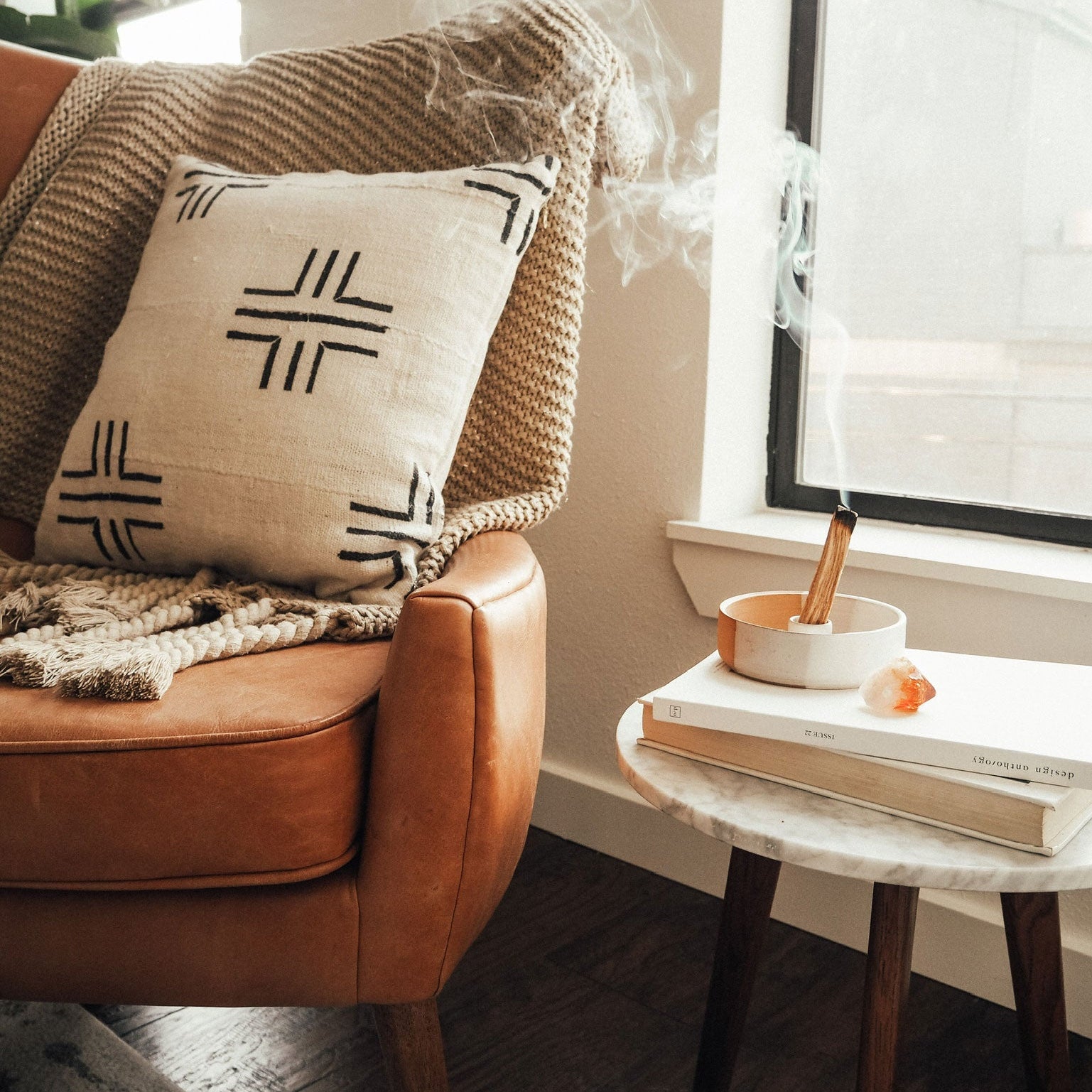 Brown leather armchair with a patterned pillow and a small table with books and a candle next to a window.