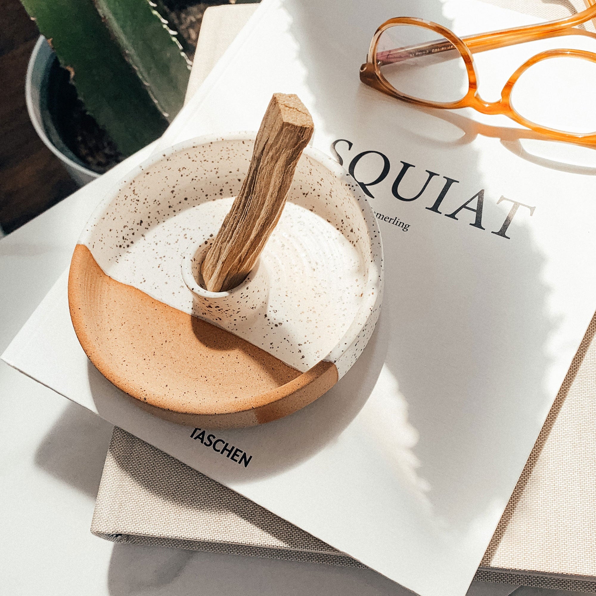Small ceramic bowl with wooden handle on a magazine with glasses and plant in the background