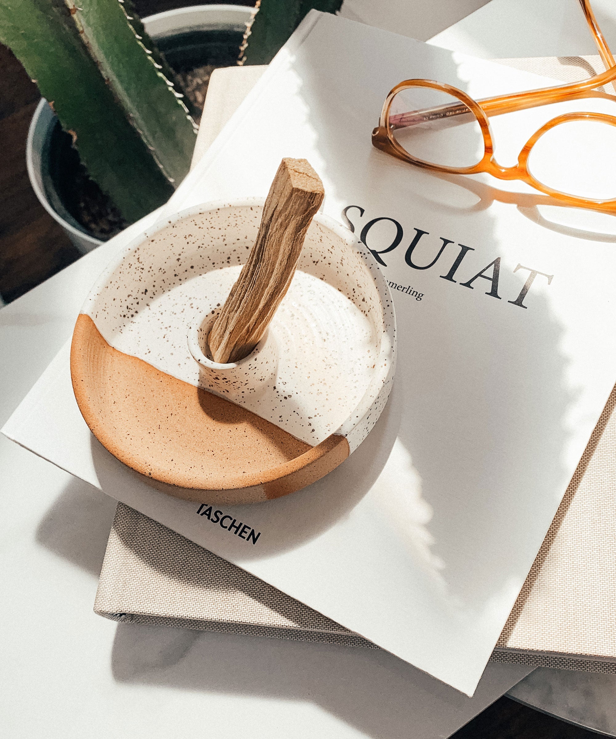 Small ceramic bowl with wooden handle on a magazine with glasses and plant in the background