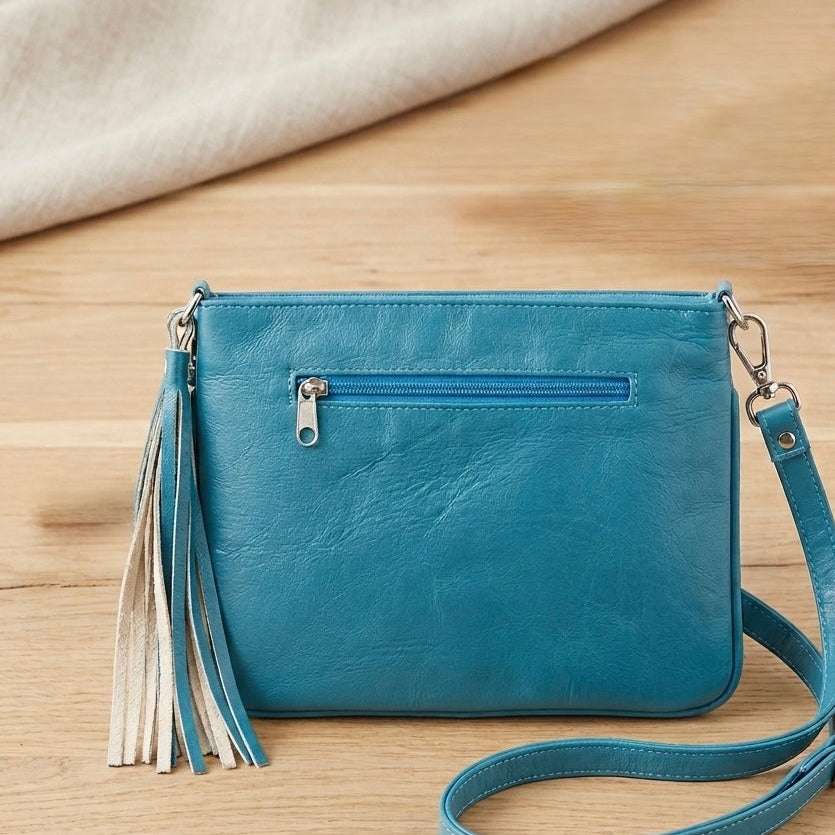 OldTribes A teal blue faux-leather crossbody clutch bag with a front zip pocket, decorative tassel, and matching shoulder strap, displayed on a light wood surface with a cream fabric background.