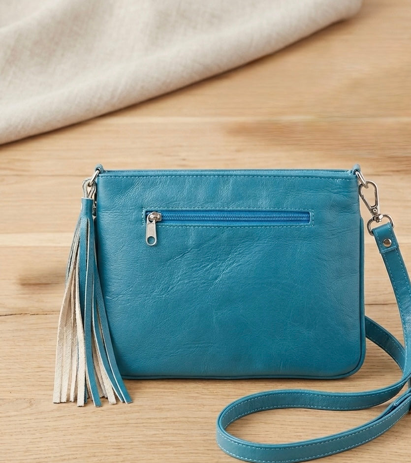 OldTribes A teal blue faux-leather crossbody clutch bag with a front zip pocket, decorative tassel, and matching shoulder strap, displayed on a light wood surface with a cream fabric background.