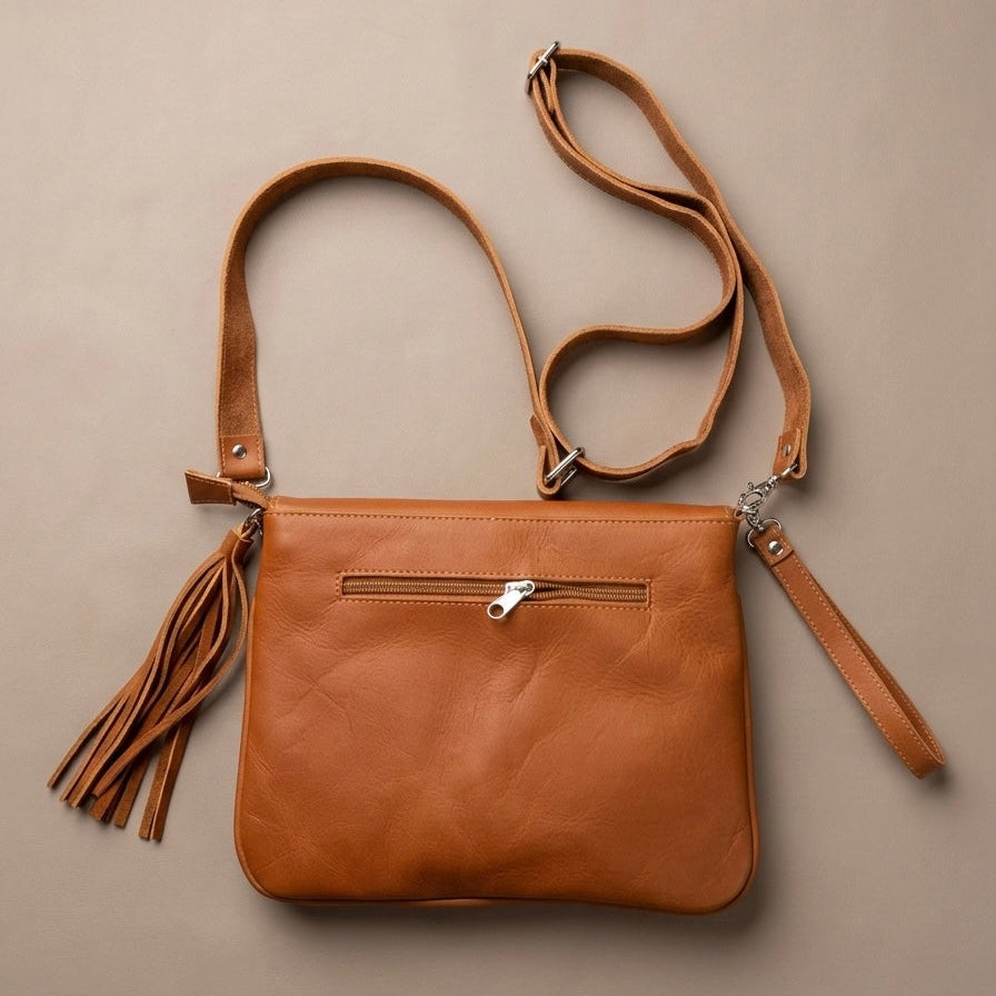 OldTribes A tan leather crossbody bag with a front zip pocket, a long adjustable shoulder strap, a wristlet strap, and a hanging tassel, displayed flat on a neutral beige background.