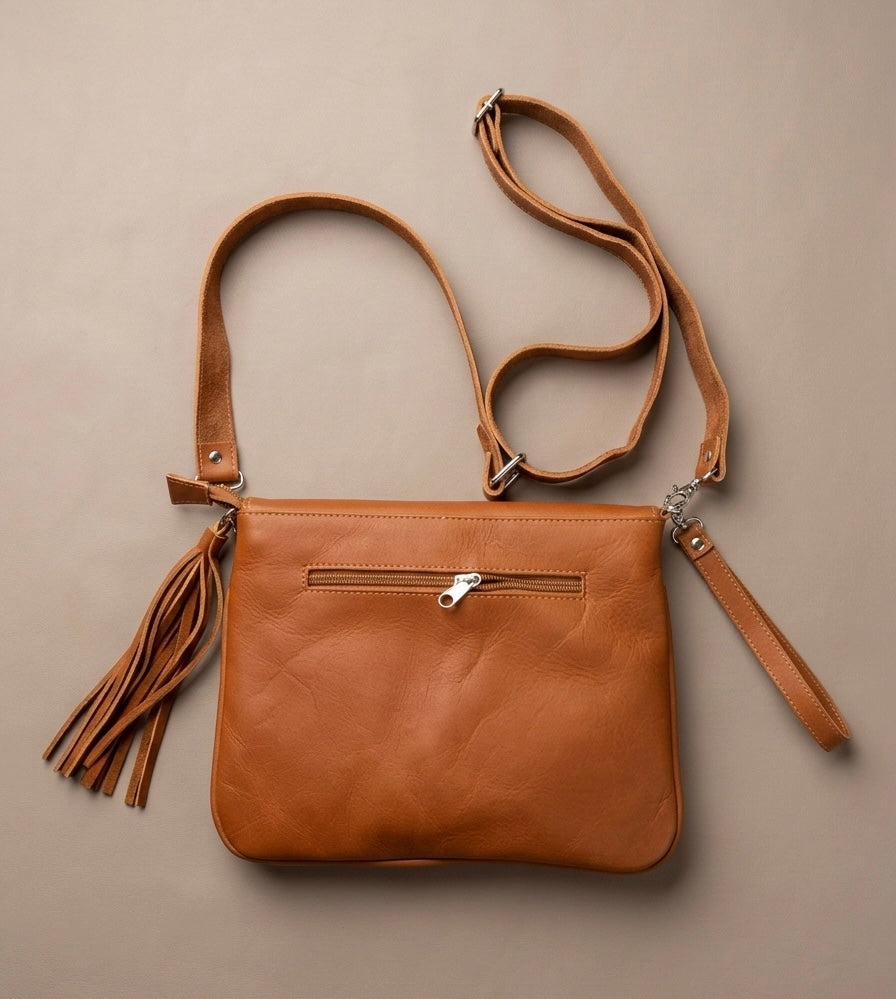 OldTribes A tan leather crossbody bag with a front zip pocket, a long adjustable shoulder strap, a wristlet strap, and a hanging tassel, displayed flat on a neutral beige background.
