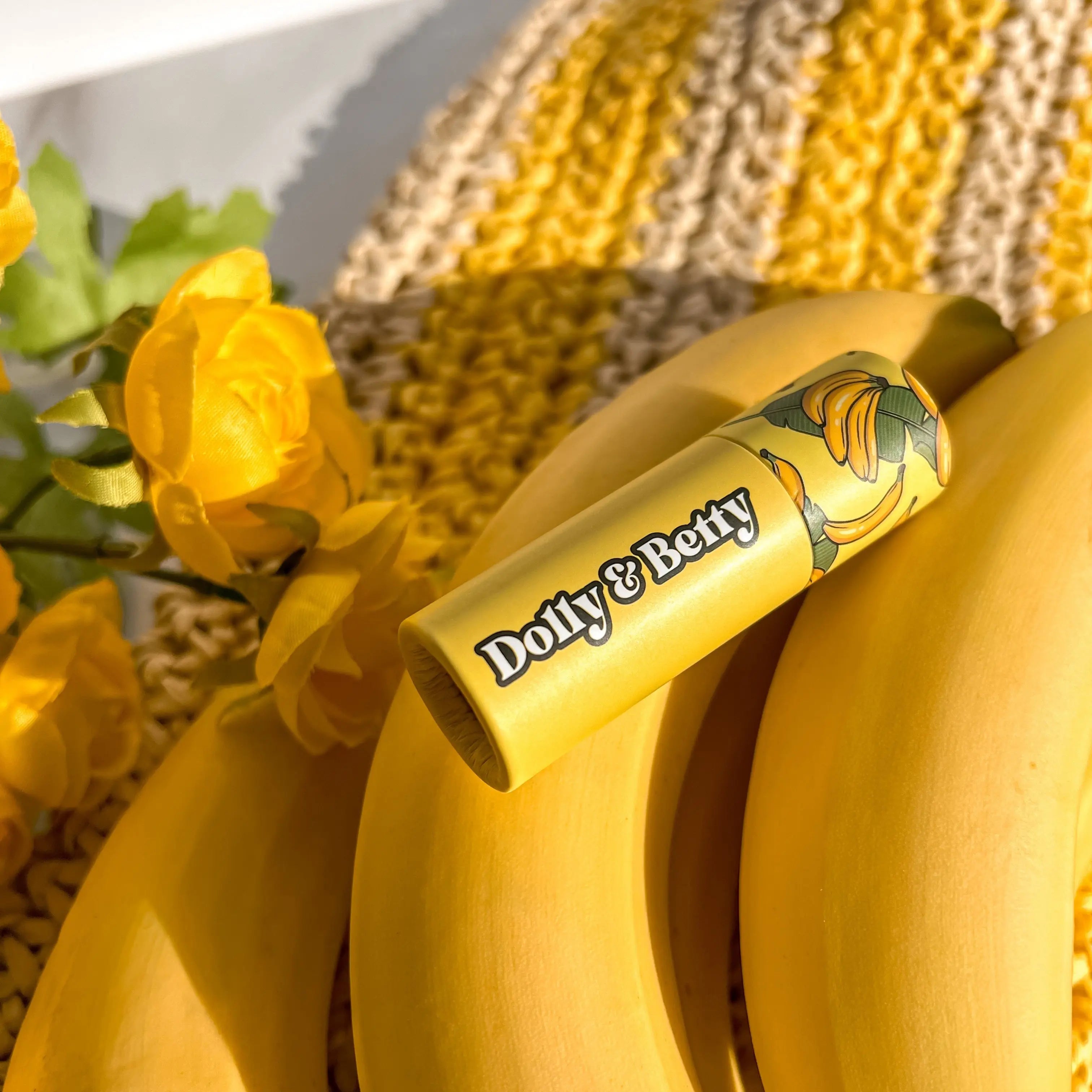 Bananas with a yellow lip balm labeled 'Dolly & Betty' against a yellow floral and straw hat background.