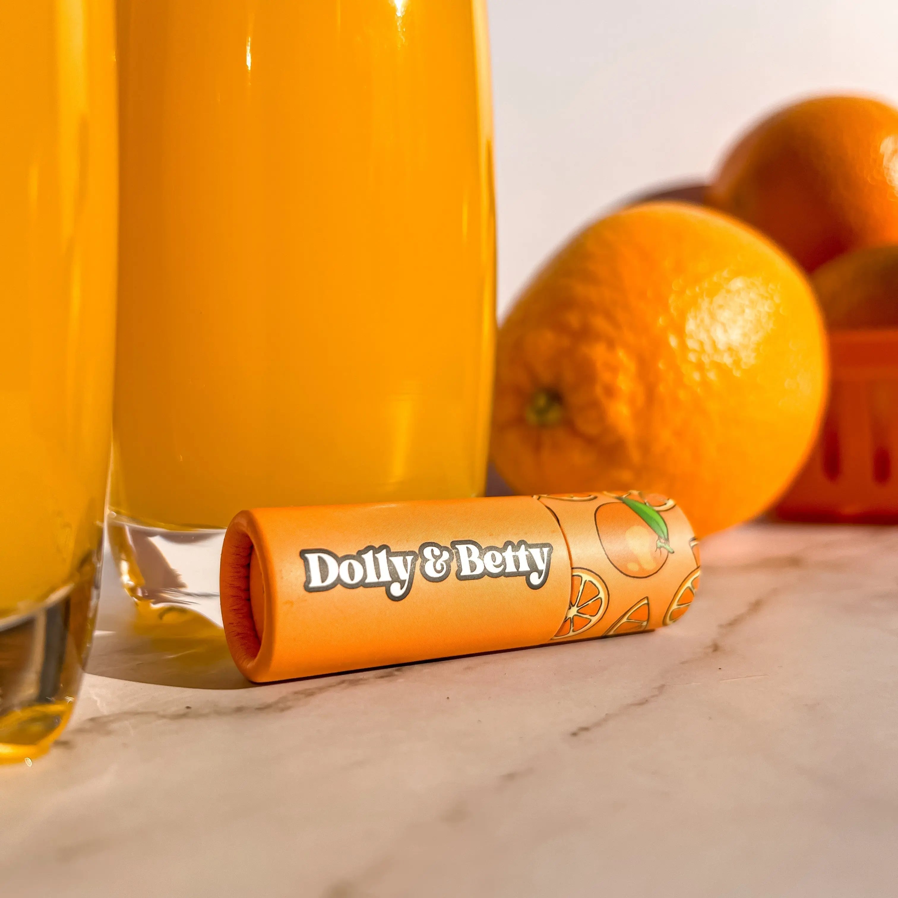 Two glasses of orange juice with a tube of Dolly & Betty on a marble surface with oranges in the background.