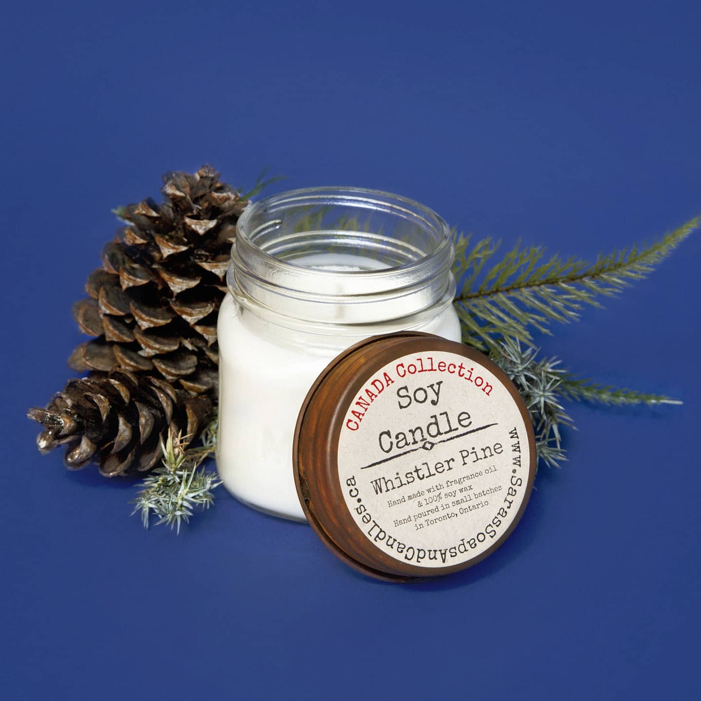 Soy candle in a jar with a wooden lid on a blue background, surrounded by pine cones and leaves.
