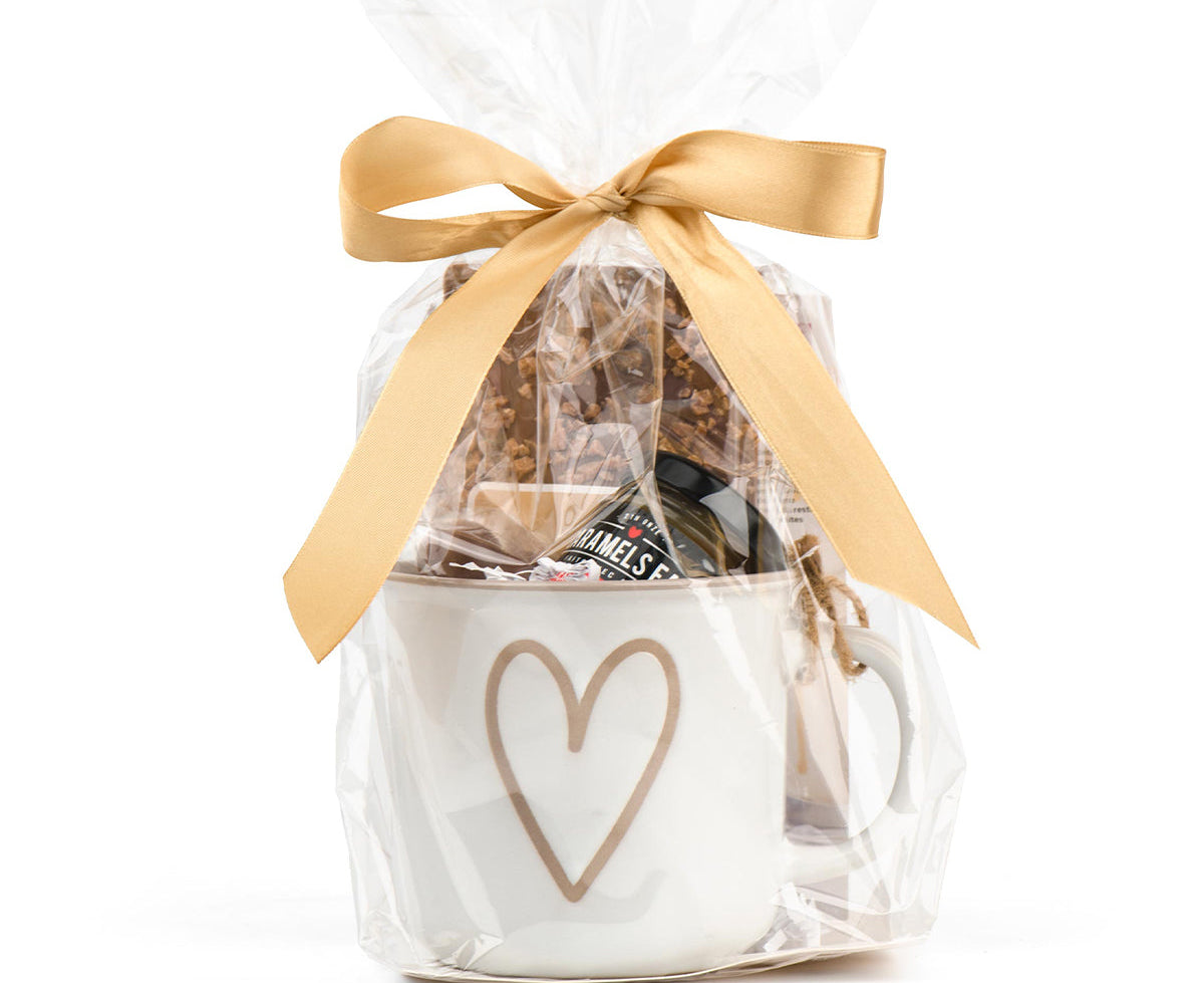 Gift basket with a white bag featuring a heart design, filled with snacks, and tied with a gold ribbon on a white background.
