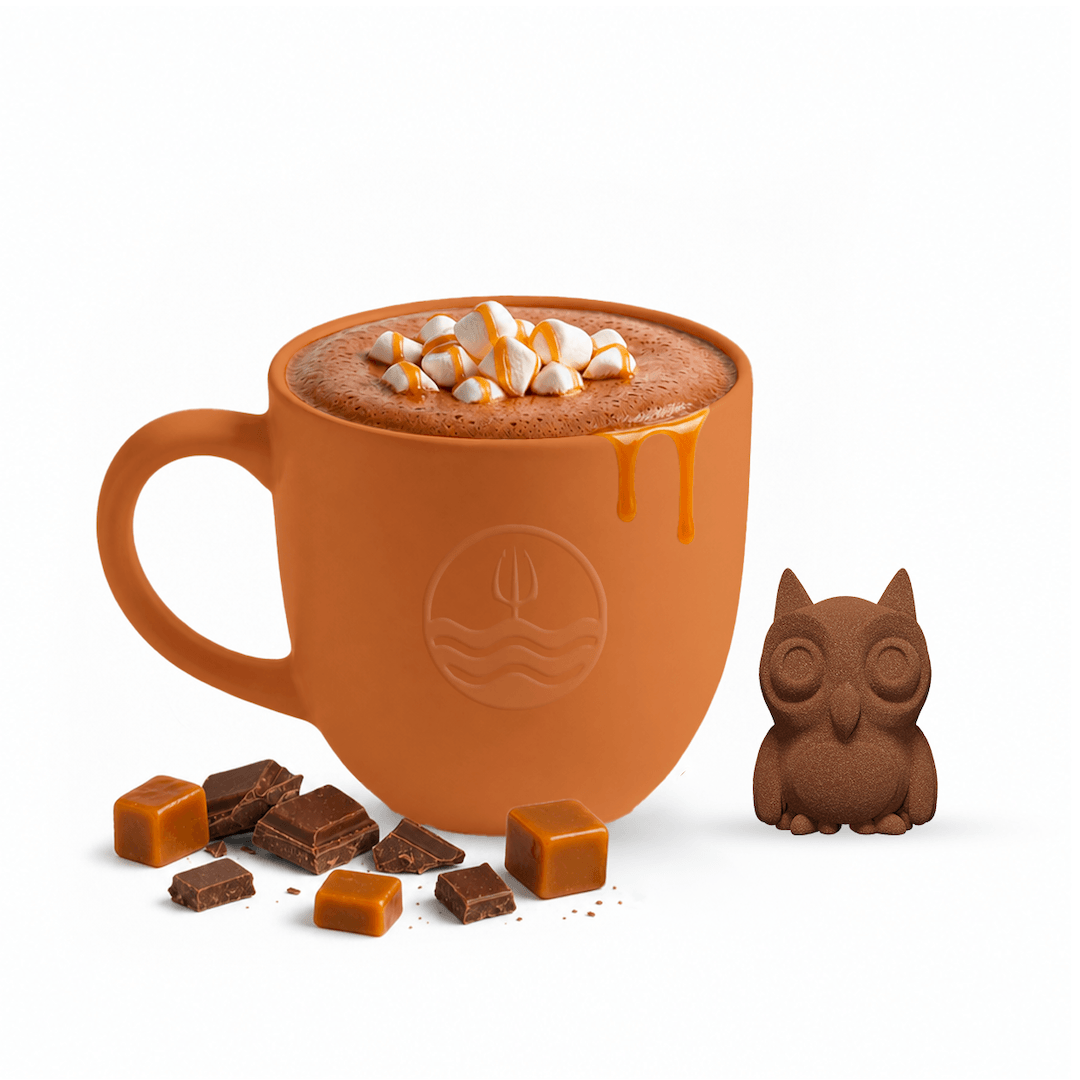 An orange ceramic mug filled with hot chocolate topped with marshmallows and caramel drizzle, styled with scattered chocolate pieces and caramel cubes on a white background, alongside a small owl-shaped chocolate figure.