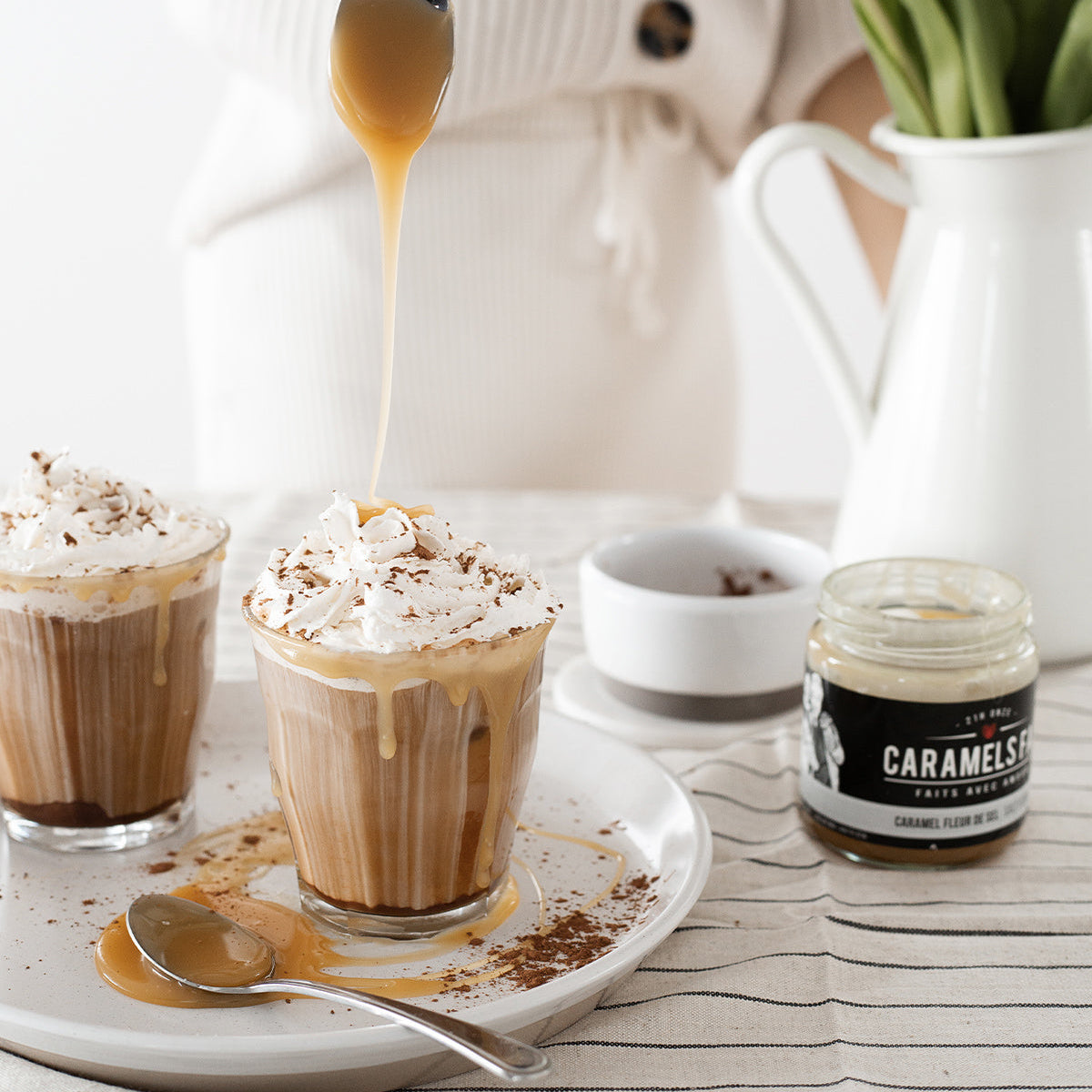 Two glasses of a creamy beverage with whipped cream and caramel sauce, a jar of caramel sauce, and a person in the background.