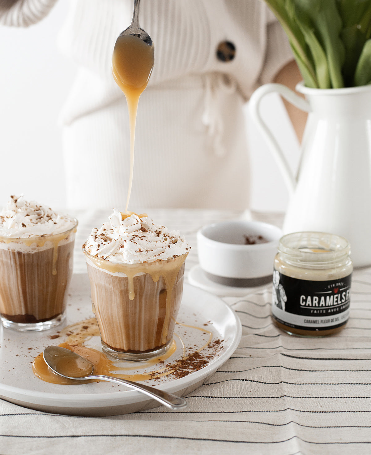 Two glasses of a creamy beverage with whipped cream and caramel sauce, a jar of caramel sauce, and a person in the background.