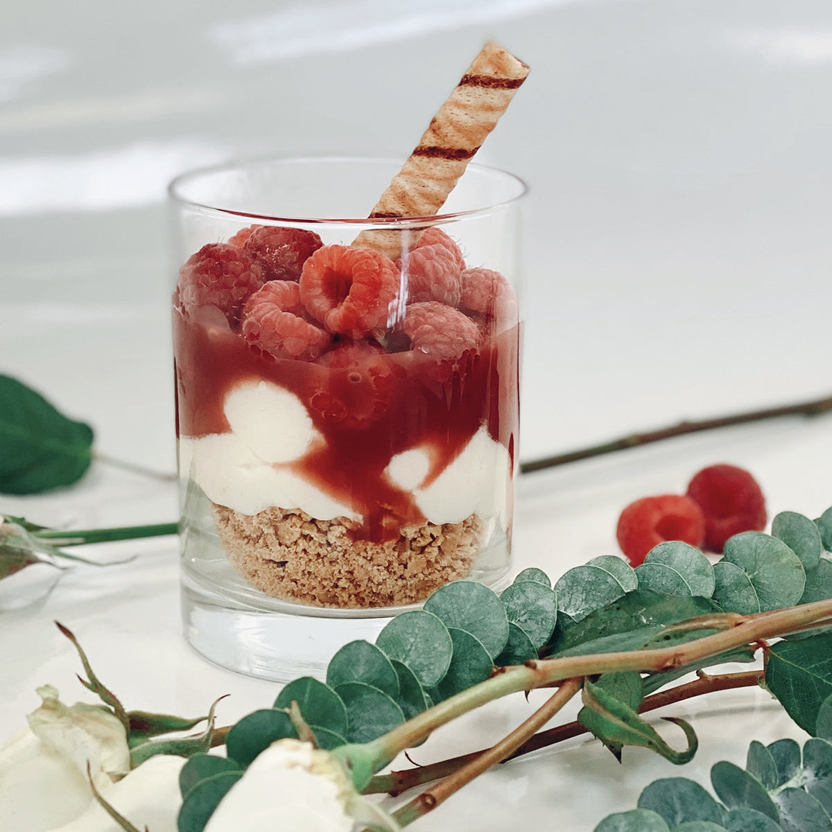 Glass dessert with raspberries, cream, and a wafer, surrounded by green leaves and white flowers on a light background.