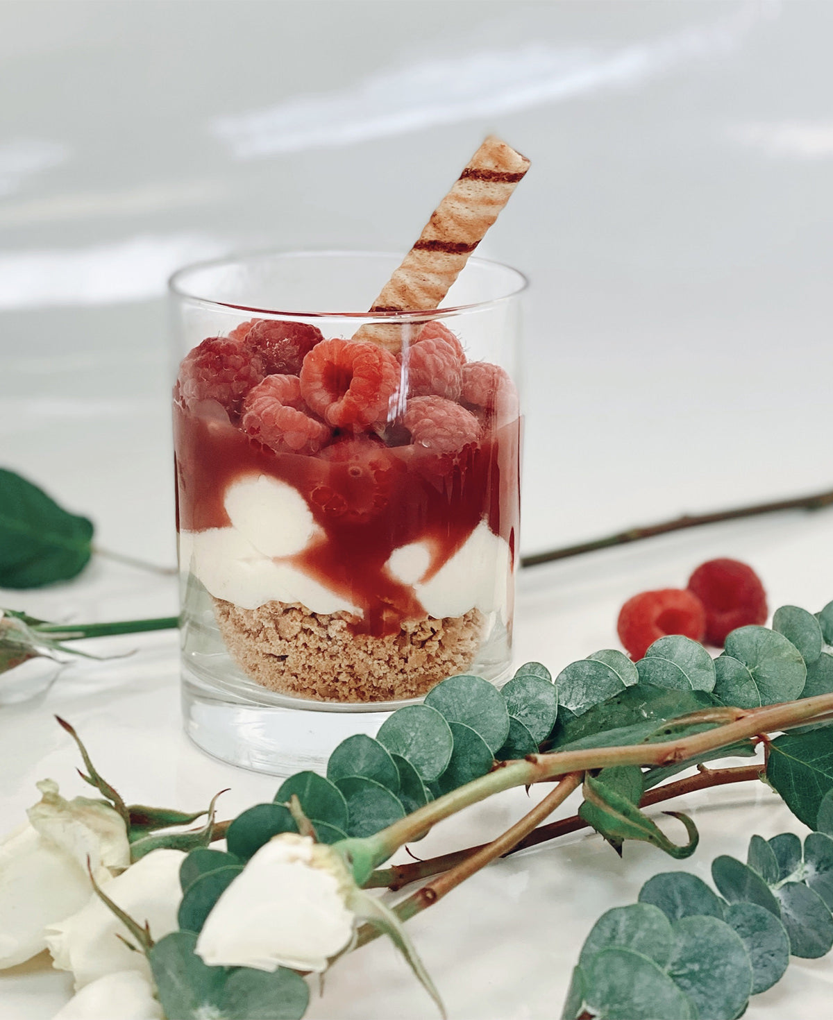 Glass dessert with raspberries, cream, and a wafer, surrounded by green leaves and white flowers on a light background.
